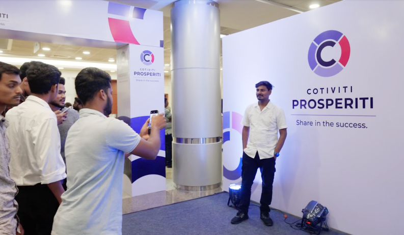 Shows a man taking a picture of another man standing in front of a banner that says Cotiviti Prosperiti: Share in the success.